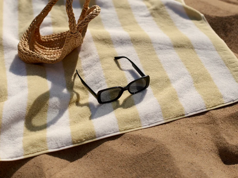 Pasiasty ręcznik plażowy rozłożony na piasku obok słomkowej torby i okularów